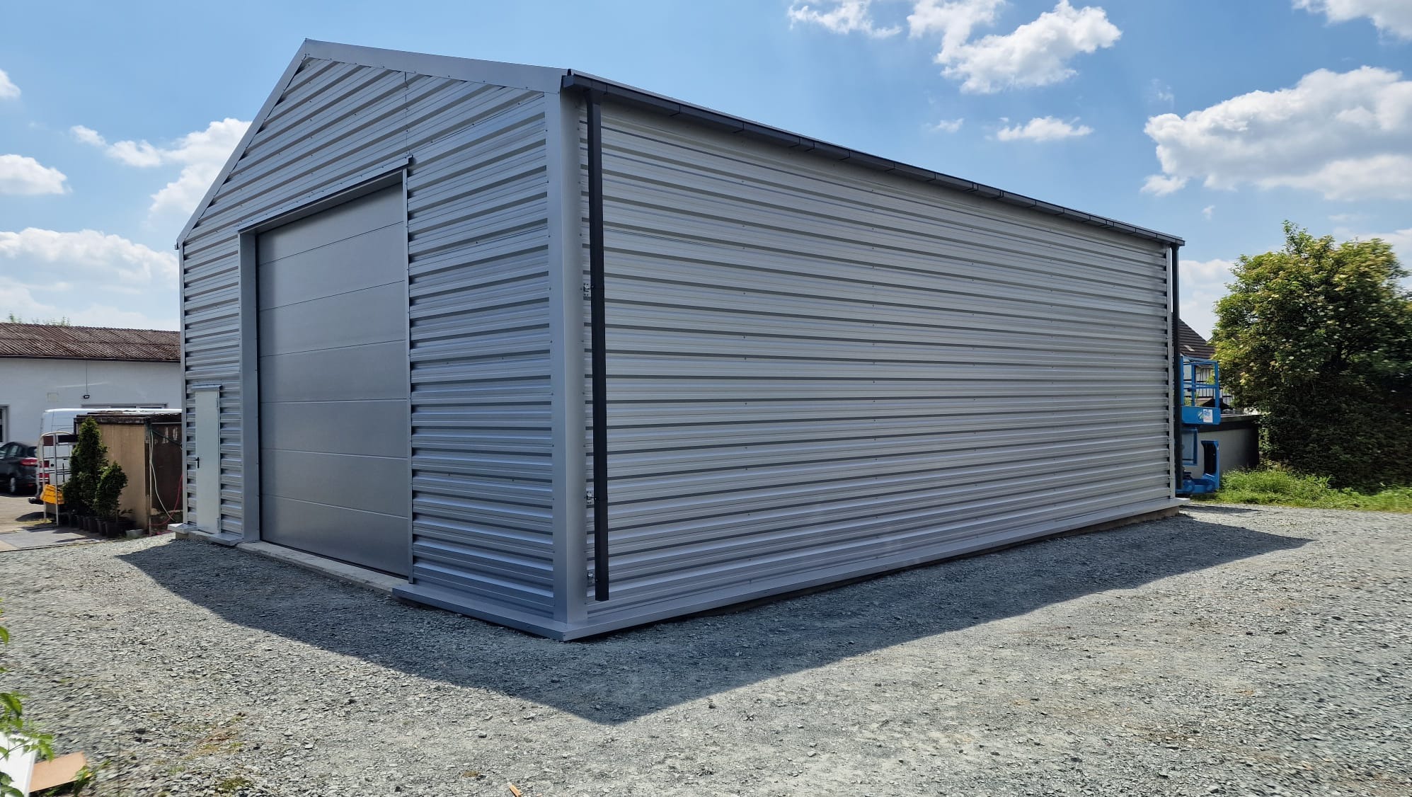 Gotowa hala magazynowa z szarej blachy trapezowej z przesuwną bramą, na żwirowym podłożu, błękitne niebo z chmurami w tle.