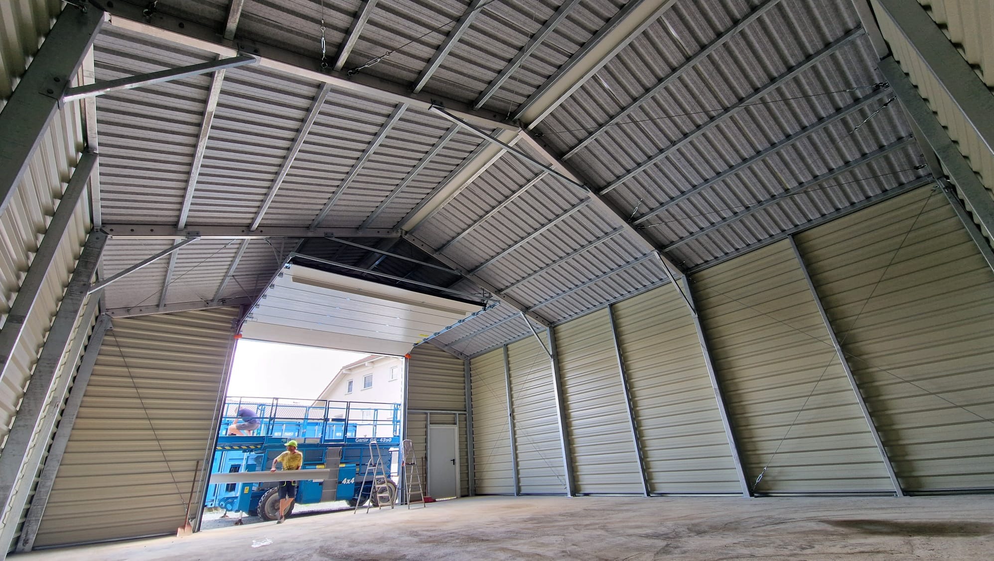 Wnętrze hali stalowej w trakcie montażu, widoczny pracownik na podnośniku koszowym i konstrukcja dachu z blachy trapezowej.