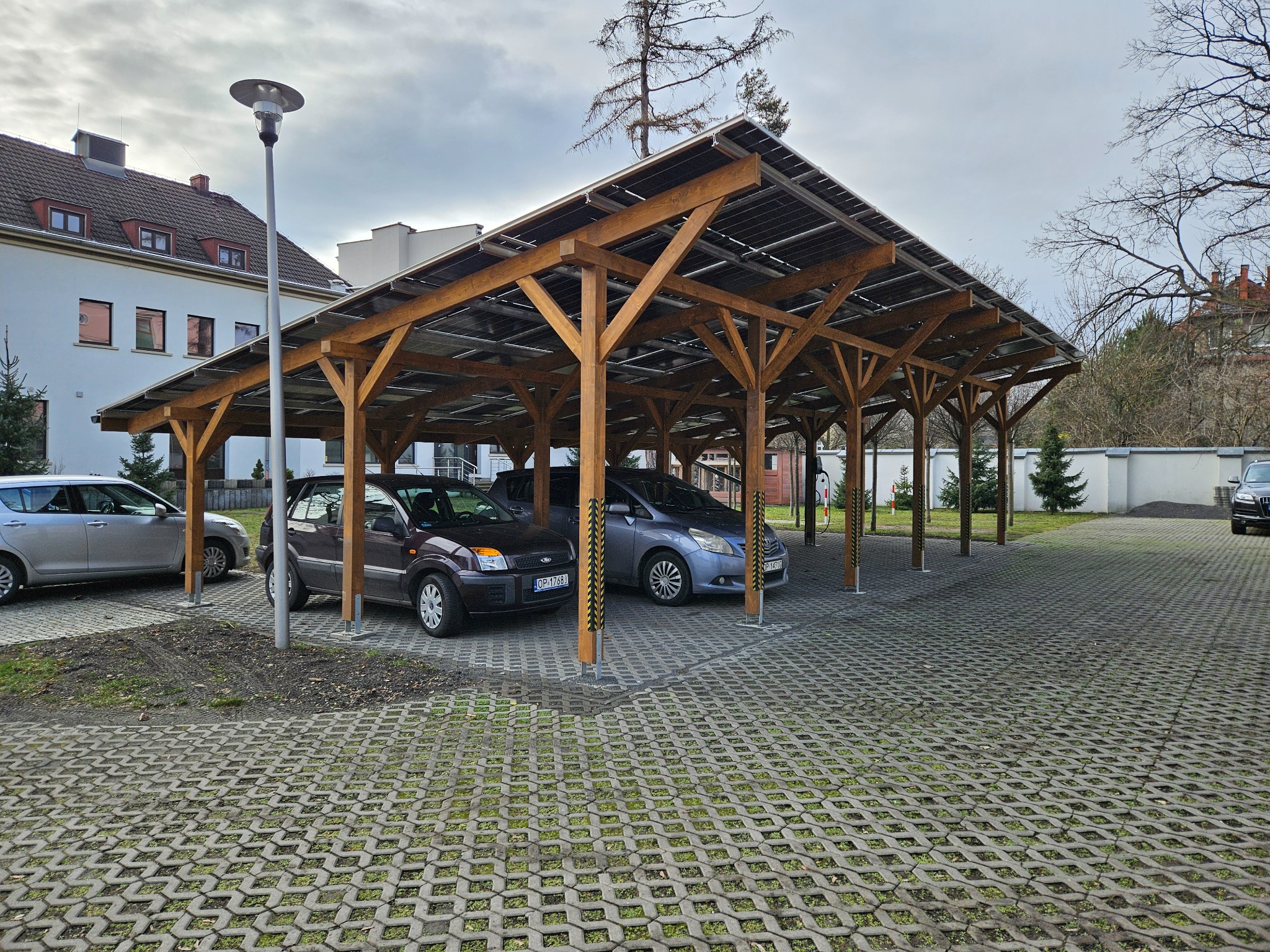 Zadaszony parking z drewnianą konstrukcją i panelami słonecznymi, z zaparkowanymi samochodami i kostką brukową z widoczną zielenią.