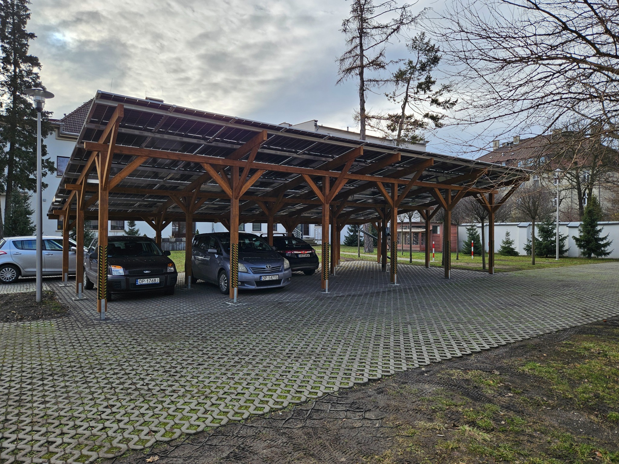 Zadaszony parking z drewnianą konstrukcją i panelami fotowoltaicznymi, z zaparkowanymi samochodami i ażurową nawierzchnią z kostki betonowej.