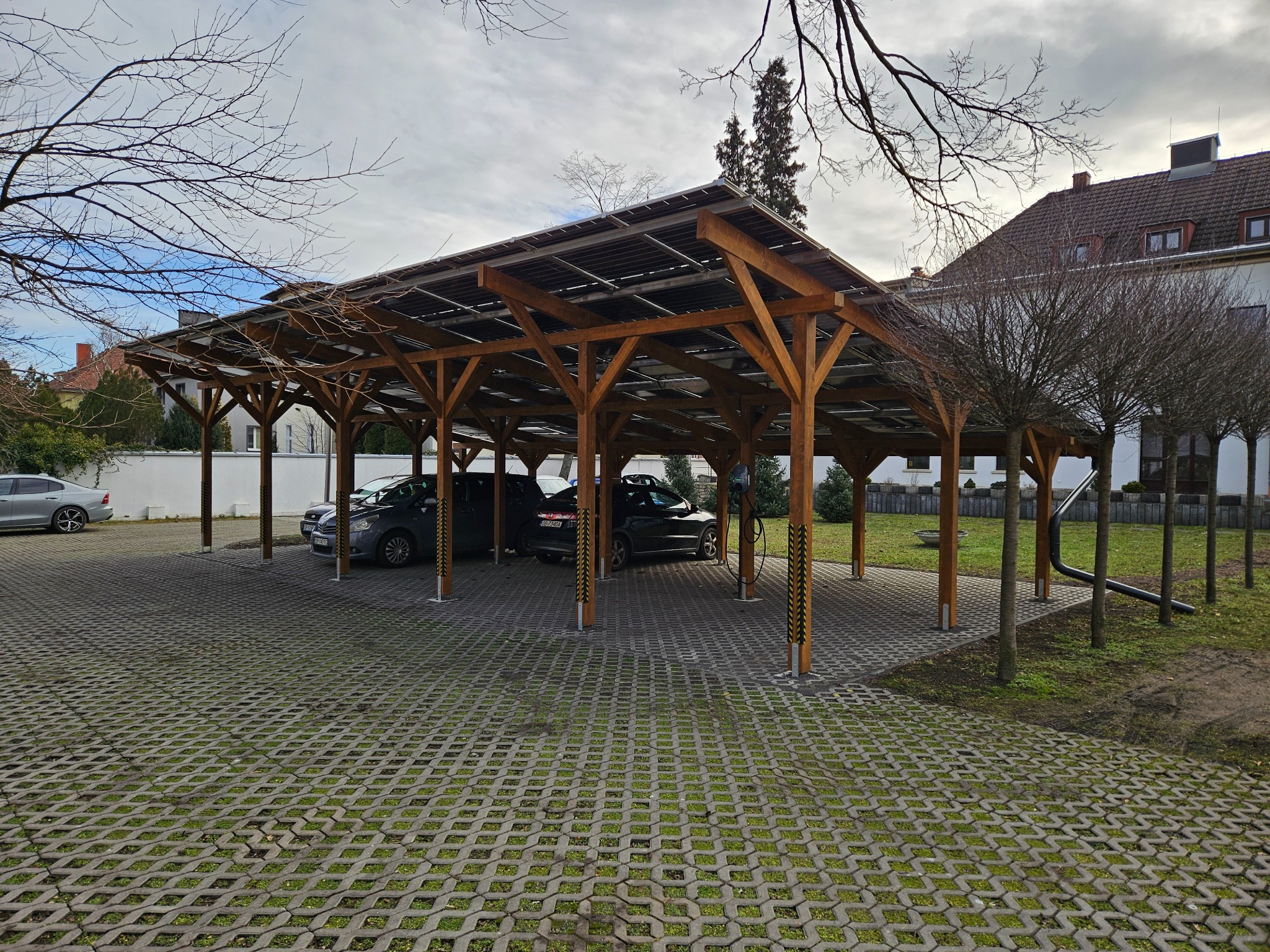 Drewniana wiata parkingowa z panelami fotowoltaicznymi na dachu, z zaparkowanymi samochodami na ażurowym parkingu z zielenią, w tle budynek.