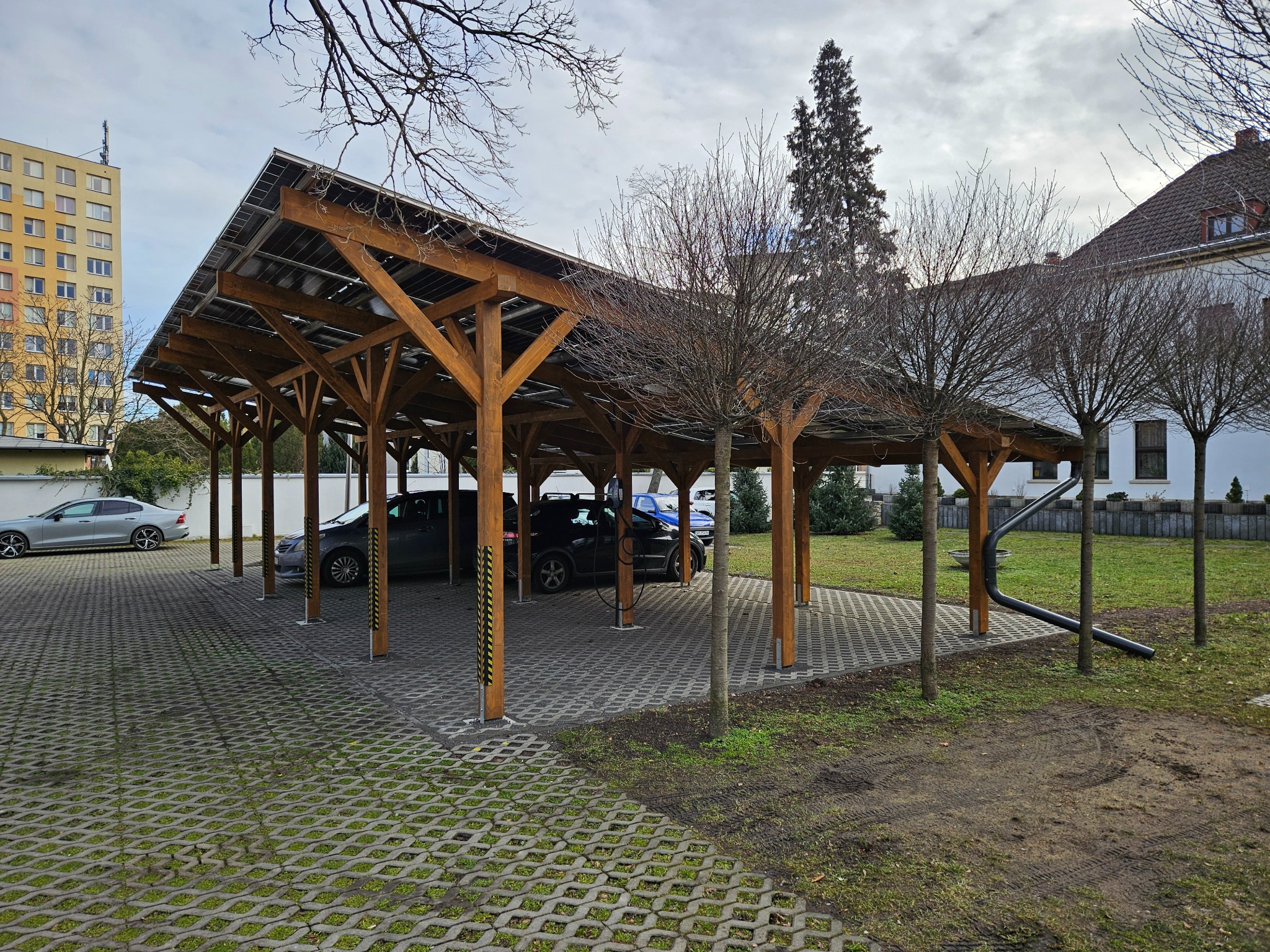 Drewniana wiata parkingowa z panelami słonecznymi, stojące pod nią samochody, ażurowa nawierzchnia z zielenią w tle budynek mieszkalny.