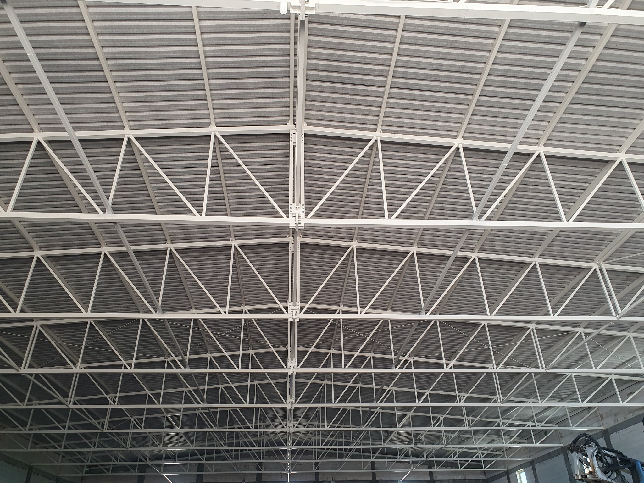 Wnętrze hali produkcyjnej z widoczną białą konstrukcją stalową dachu i szarą blachą trapezową, sfotografowane z dołu.