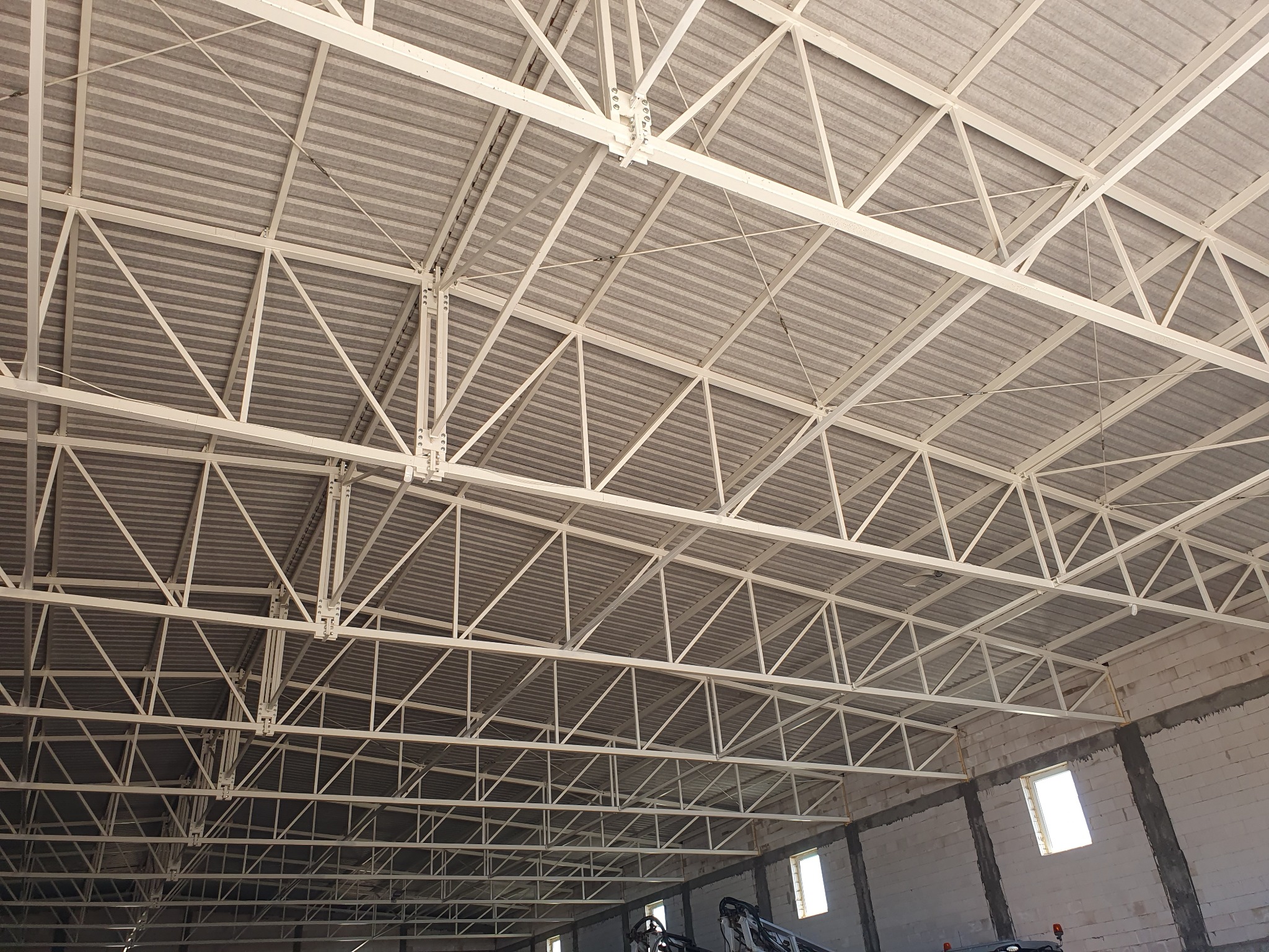 Wnętrze hali w trakcie budowy, widok na stalową konstrukcję dachu z szarego pokrycia, ściany z białych bloczków z oknami.