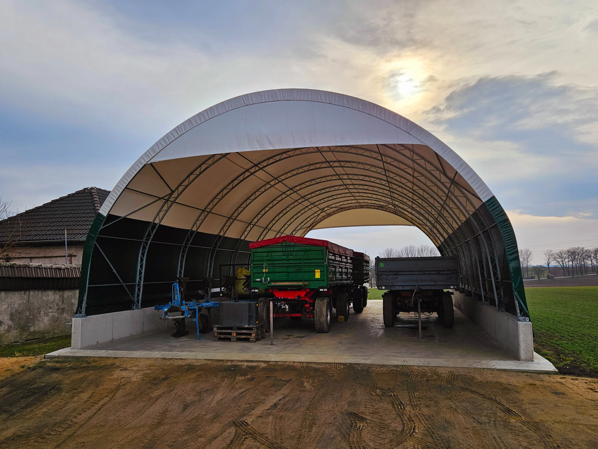 Stalowa hala łukowa z plandeką, wewnątrz zaparkowane przyczepy rolnicze, na zewnątrz pole uprawne i budynek gospodarczy.
