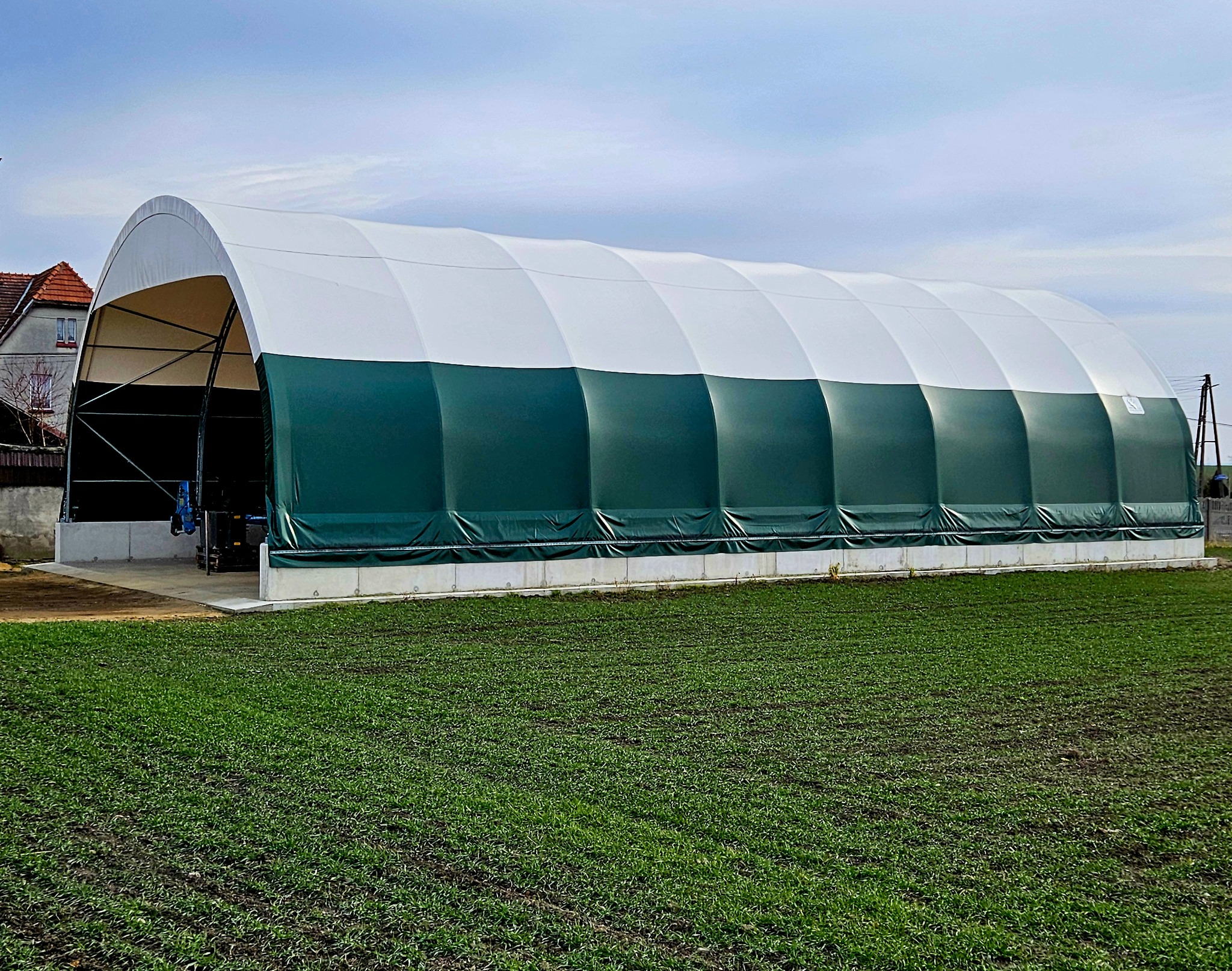 Zielono-biała hala namiotowa z metalową konstrukcją w Opolu, widoczna z zewnątrz na tle zielonego pola i zabudowań mieszkalnych.