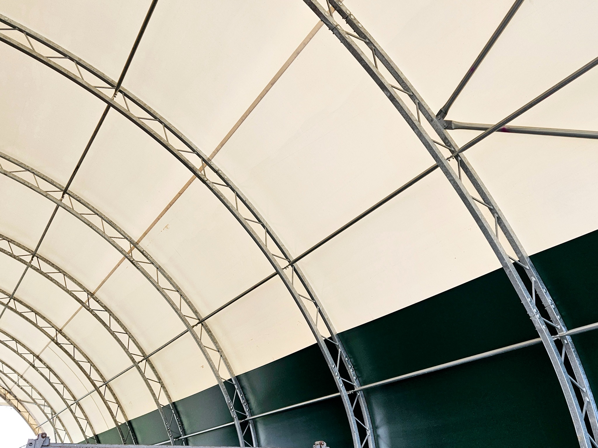 Wnętrze hali namiotowej z widoczną konstrukcją stalową łukową i białym poszyciem dachowym oraz ciemnozielonymi ścianami bocznymi.