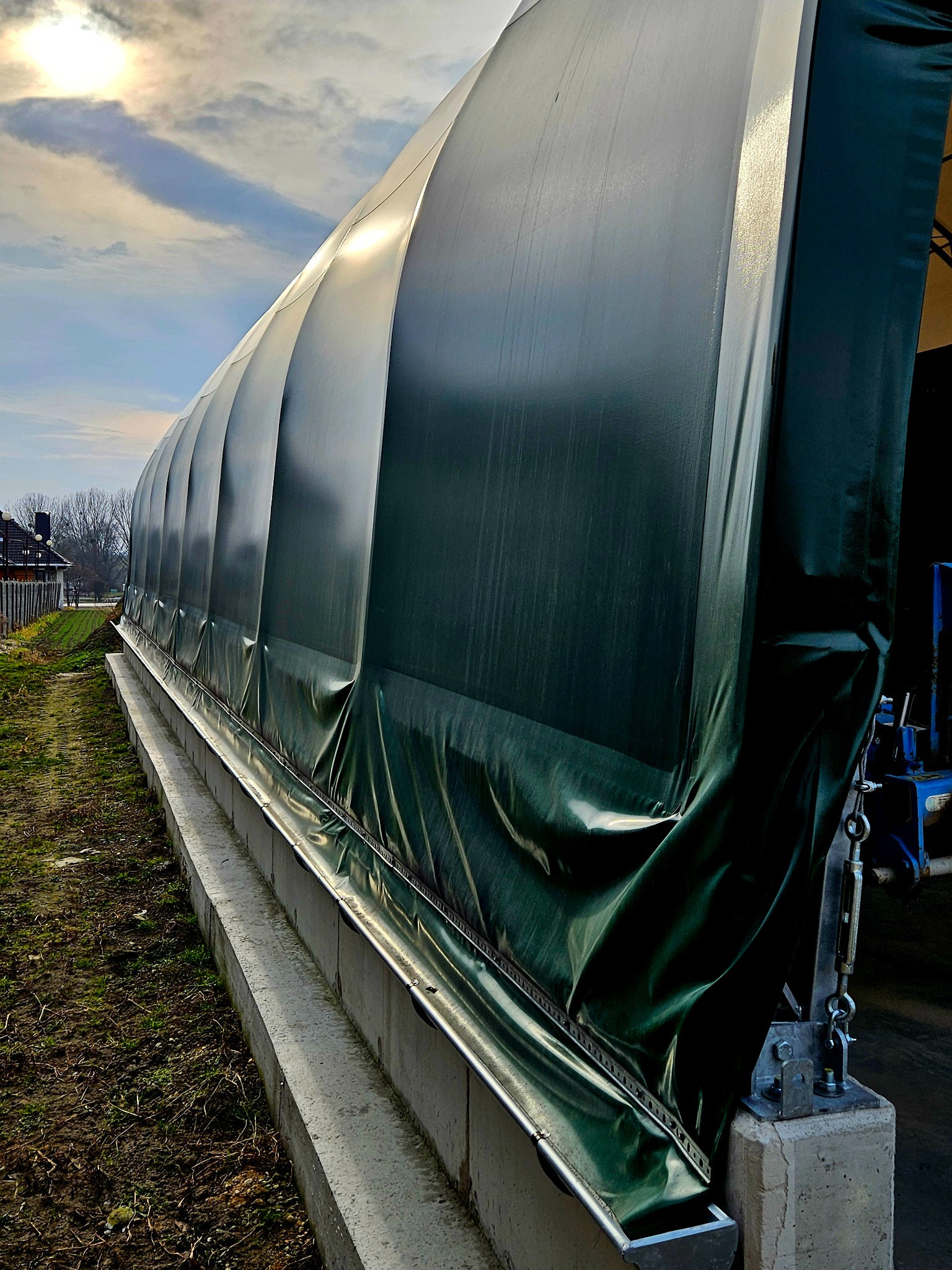 Zielona, plandekowa hala namiotowa z metalową konstrukcją, widok z zewnątrz, w tle błękitne niebo z chmurami.