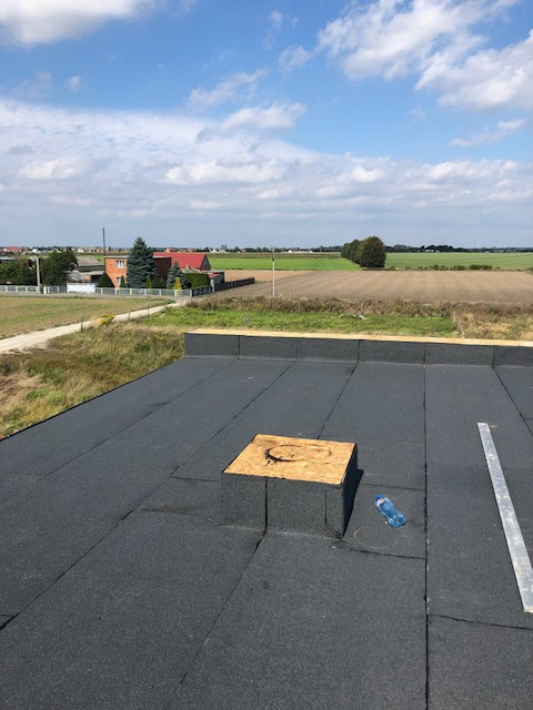 Widok z góry na czarną, płaską powierzchnię dachu z papy, z drewnianą skrzynką wentylacyjną, butelką wody i aluminiową łatą dekarską, na tle krajobrazu wiejskiego z polami i zabudowaniami...