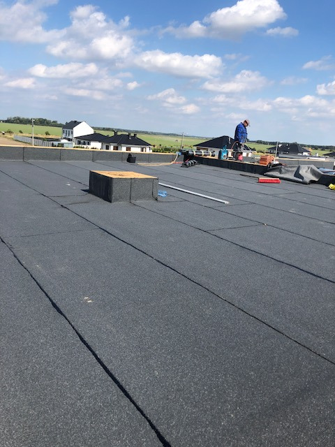 Prace na dachu: monter w niebieskim kombinezonie przy pracy na pokrytym papą dachu, widoczne narzędzia i materiały, w tle zabudowa jednorodzinna i błękitne niebo z chmurami.