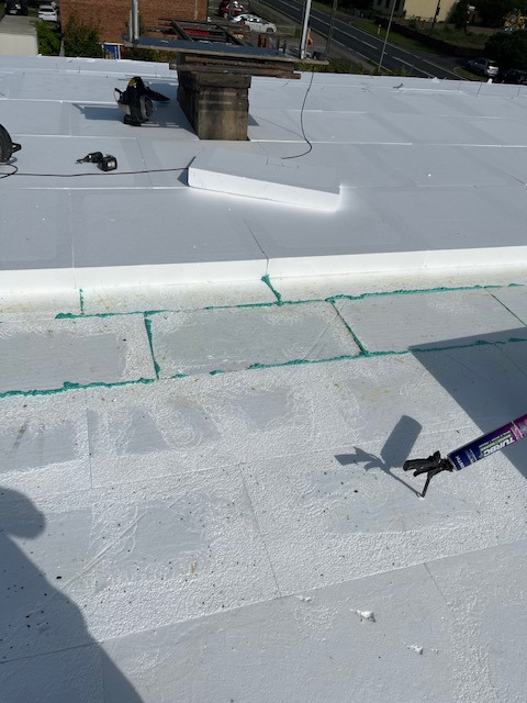 Białe płyty izolacyjne na dachu podczas montażu, widoczny aplikator kleju i narzędzia w tle, komin z betonu