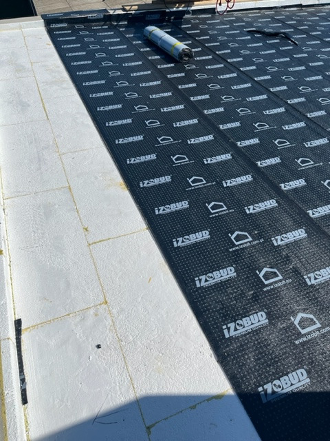 Białe płyty izolacyjne na poddaszu pokryte czarną membraną z nadrukowanym logo i napisem IZOBUD, widoczny częściowo zwinięty rulon membrany.