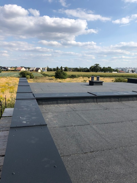 Wykończony płaski dach z papy termozgrzewalnej i obróbkami blacharskimi, widok na okoliczną zabudowę i pole w słoneczny dzień.