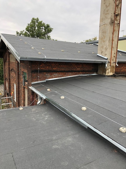 Wykończony płaski dach z papy, z metalowymi obróbkami blacharskimi i zabezpieczeniami odgromowymi, widok z góry na ceglany budynek z kominem w tle.