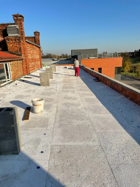 Prace na dachu: Robotnik w czerwonym kombinezonie na płaskim dachu pokrytym szarymi płytami izolacyjnymi, widoczny mur ceglany, pustaki i wiadro z materiałem izolacyjnym.