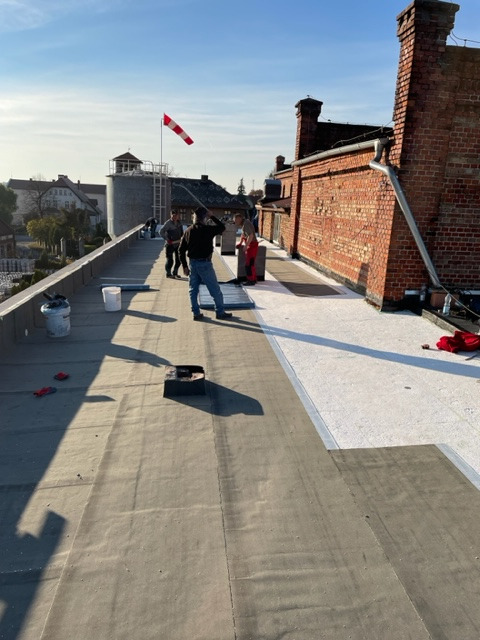 Prace na dachu: ekipa montuje izolację, widoczny wskaźnik wiatru i ceglany komin w tle.