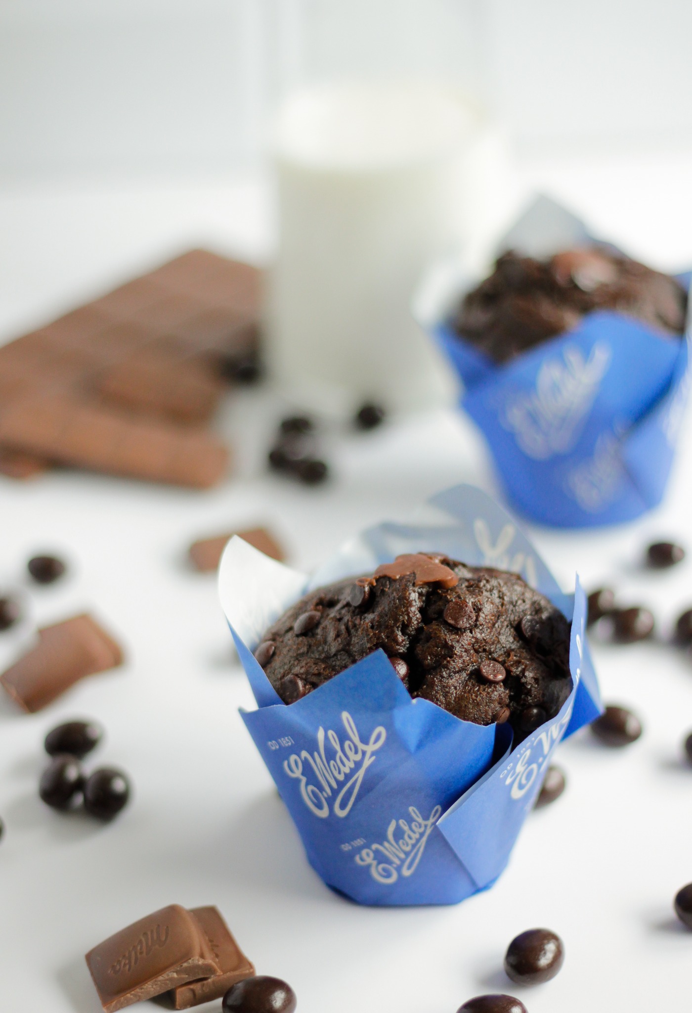 Dwa czekoladowe muffiny marki Wedel w niebieskich papilotkach, czekoladki w kształcie kulek i połamana tabliczka czekolady Milka na białym tle, w tle szklanka mleka.