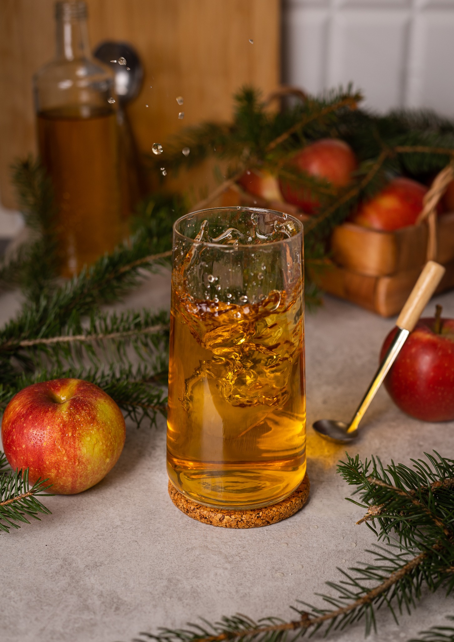 Szklanka z jabłkowym napojem na korkowej podkładce, obok jabłka, gałązki świerku i łyżeczka z drewnianą rączką, w tle butelka i koszyk z jabłkami.