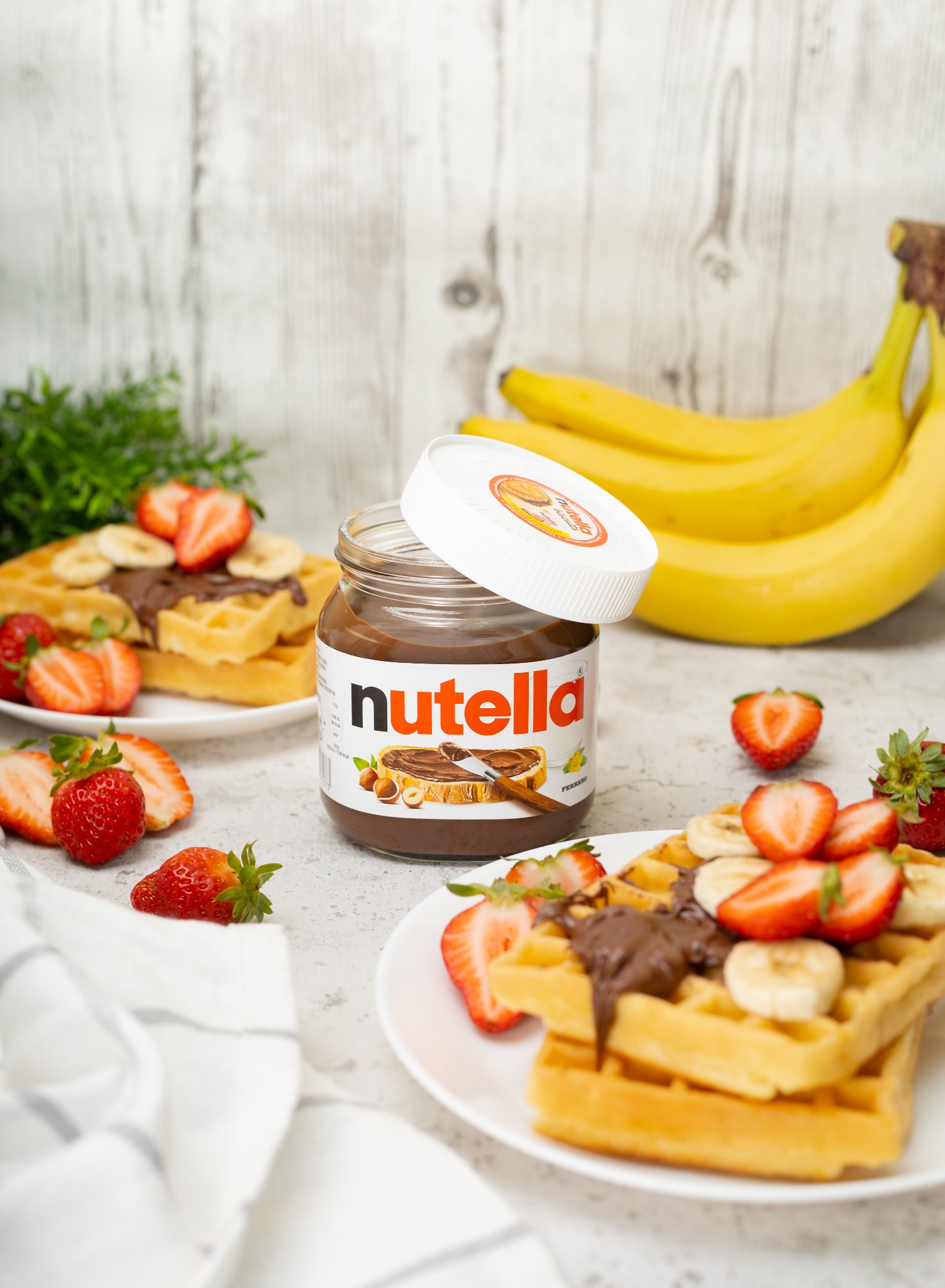 Słoik Nutelli z otwartym wieczkiem na tle wafli z bananami i truskawkami, oraz kiść bananów, ustawione na jasnym blacie.