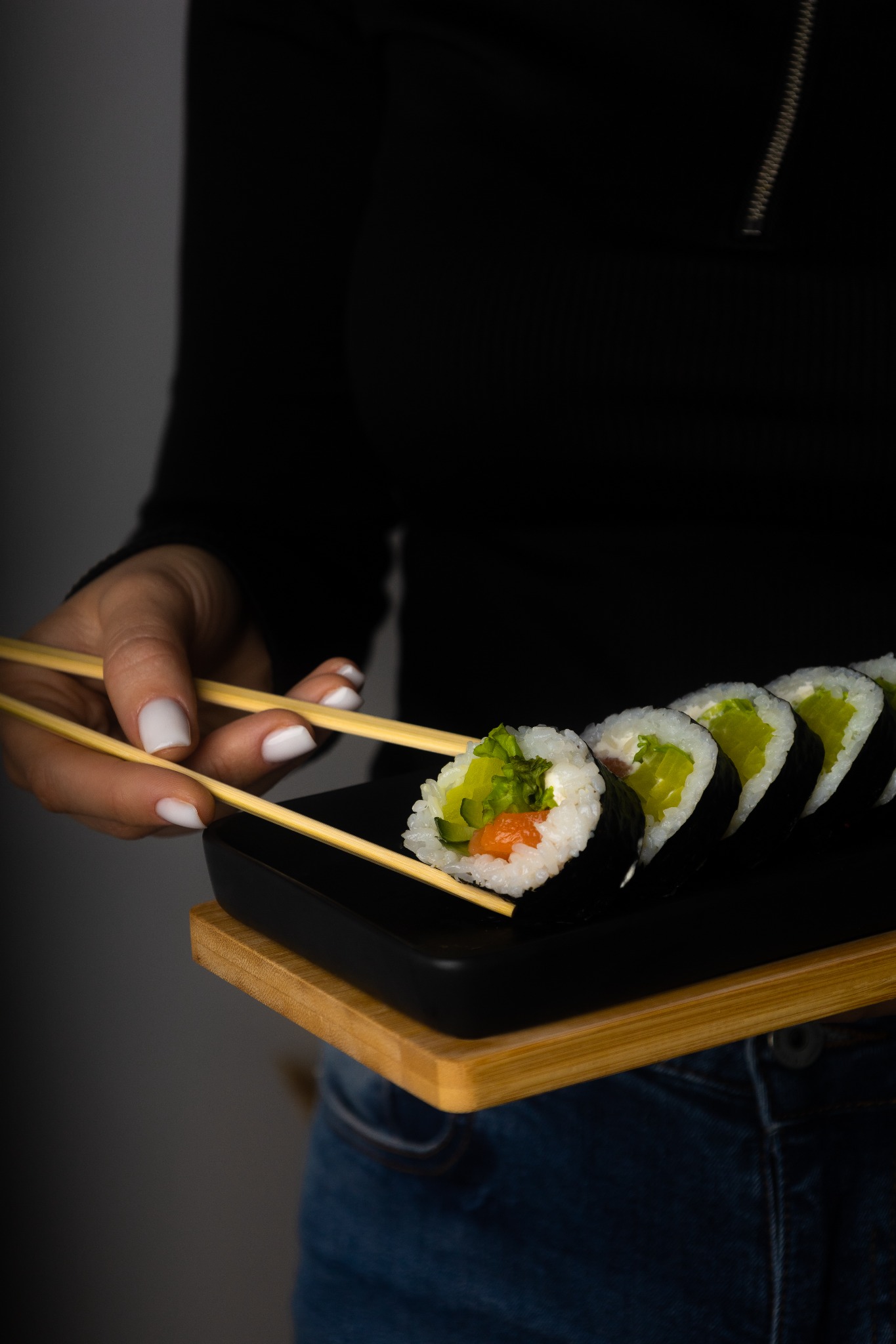 Ręka z białym manicure trzyma pałeczki nad czarnym talerzem z sushi z łososiem, sałatą i ogórkiem, na drewnianej desce.