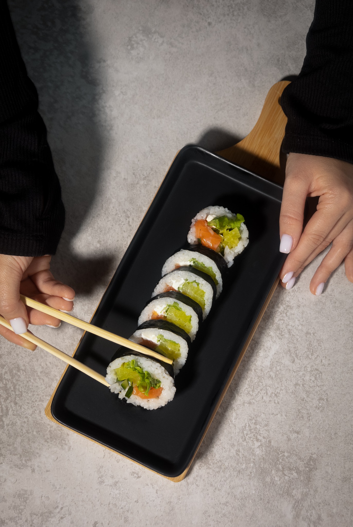 Ręce z pałeczkami podnoszą kawałek sushi z łososiem i awokado ułożonego na czarnym talerzu z bambusowym uchwytem.