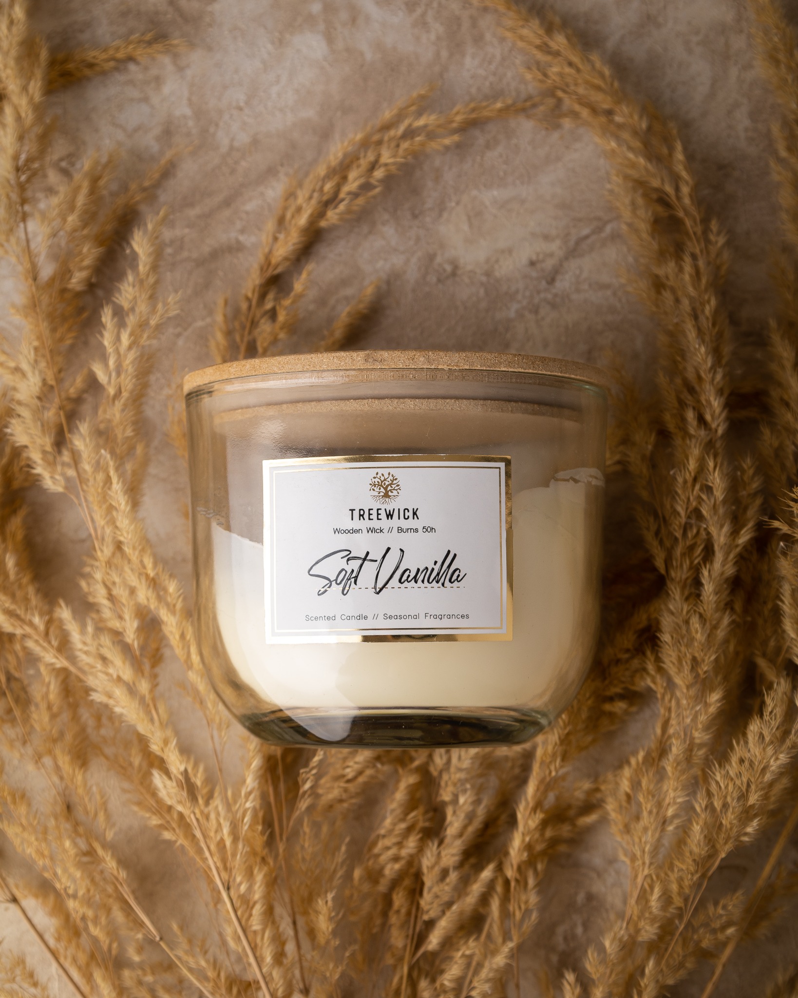 Szklany słoik z waniliową świecą zapachową Treewick, drewniany knot, etykieta z logo, otoczony suchymi trawami na beżowym tle.