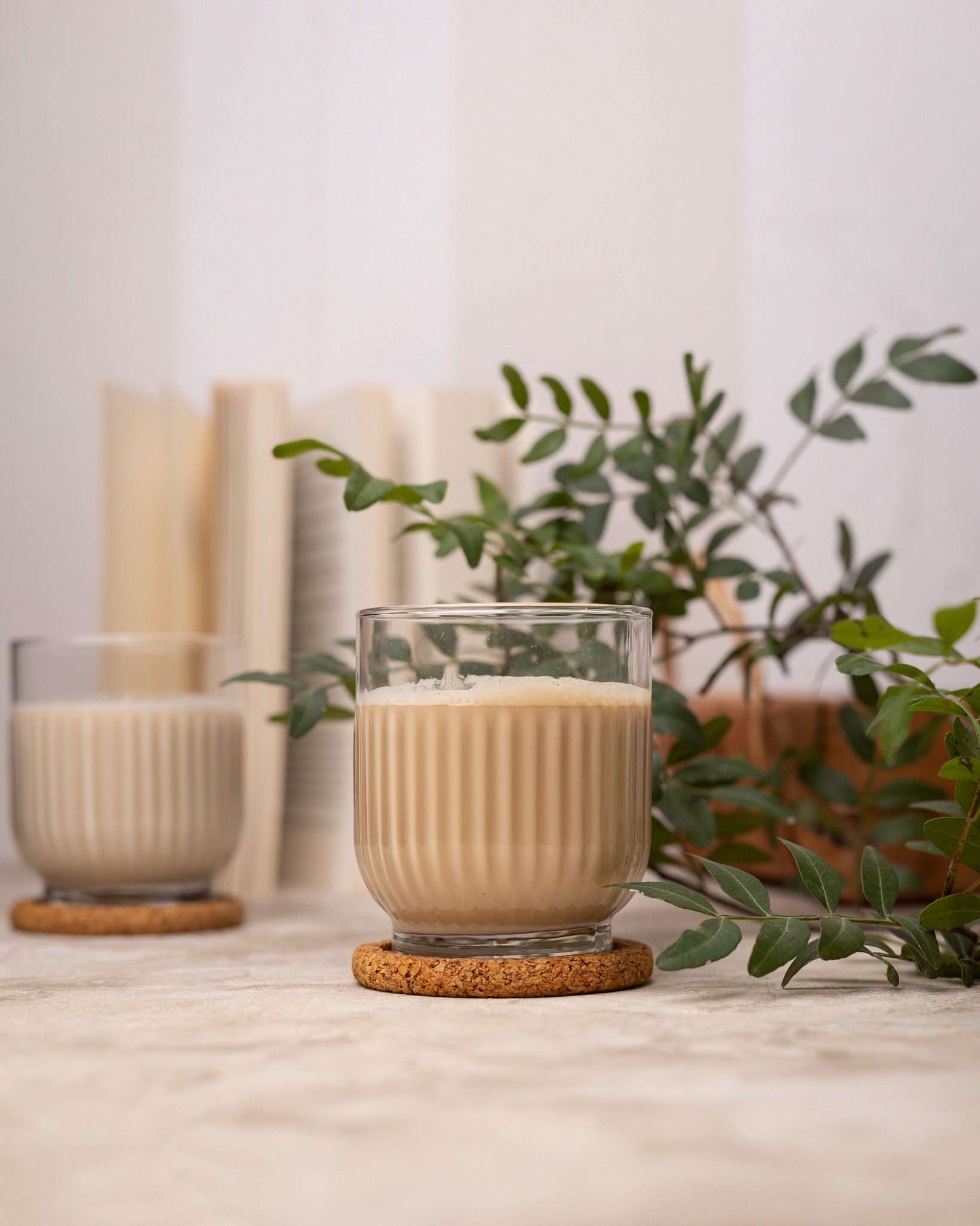 Dwa szklane kubki z beżowym napojem na korkowych podkładkach, otwarte książki i zielona gałązka w tle, miękkie światło.