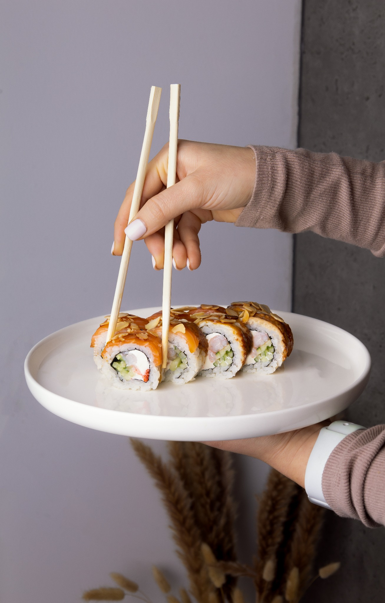Ujęcie dłoni z pałeczkami podnoszącej kawałek sushi z łososiem, ogórkiem i kremowym serkiem na białym talerzu.