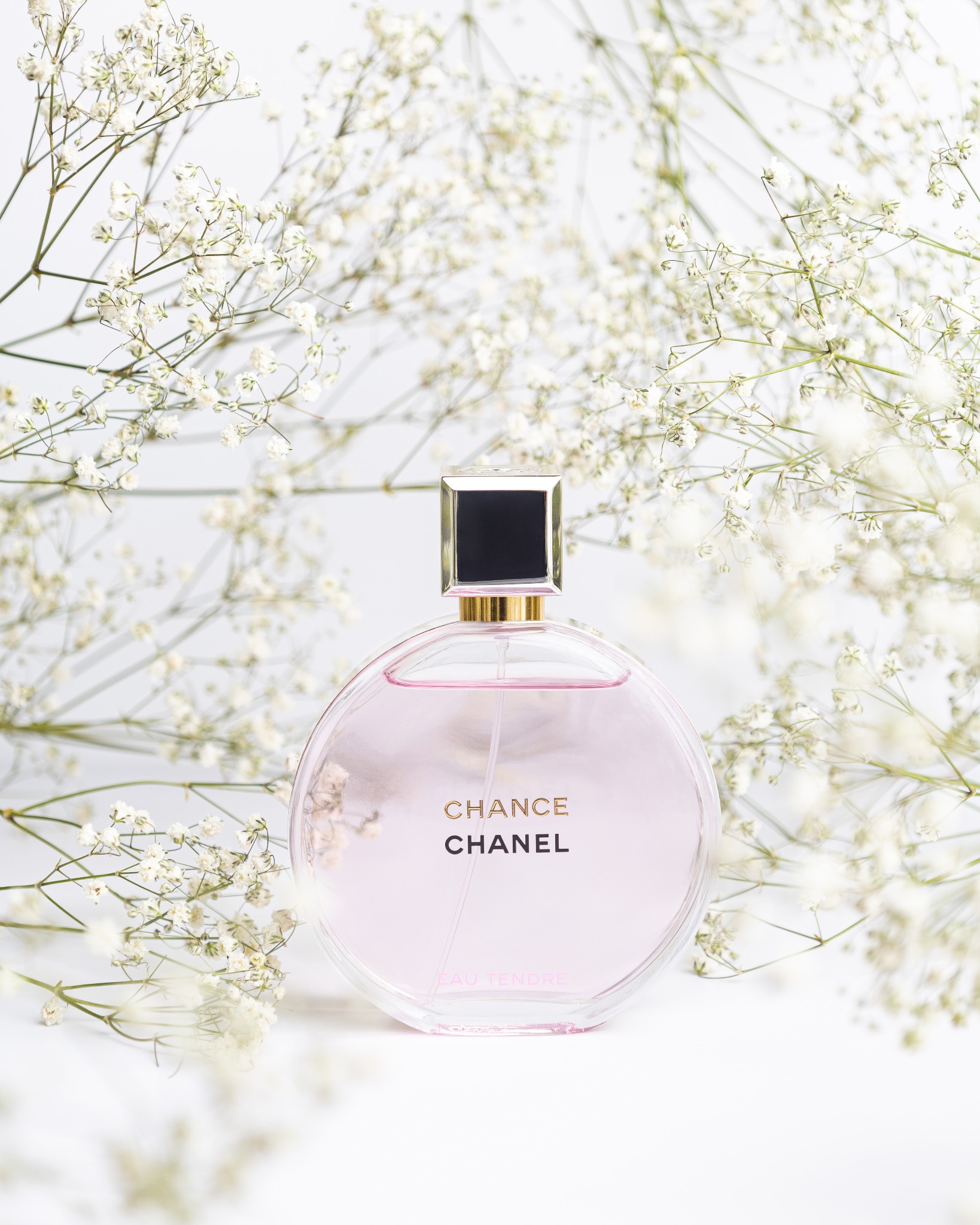 Butelka perfum Chanel Chance Eau Tendre otoczona drobnymi białymi kwiatami na białym tle, prezentacja produktu w delikatnej, subtelnej kompozycji.