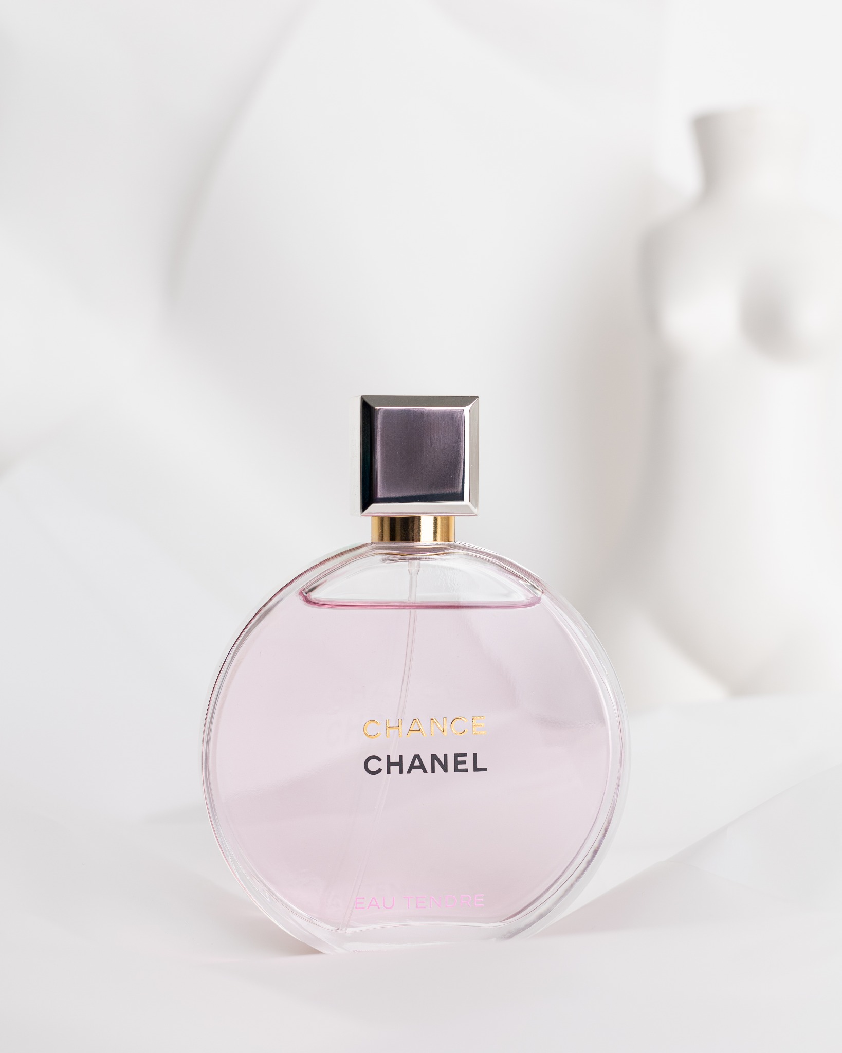 Butelka perfum Chanel Chance Eau Tendre na białym tle z miękkimi fałdami materiału i rozmytą sylwetką torsu w tle.