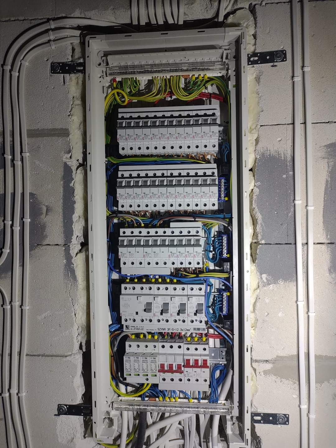 Rozdzielnica elektryczna z licznymi bezpiecznikami i okablowaniem, zamontowana na surowej ścianie z betonu komórkowego, częściowo wypełnionej pianką montażową.