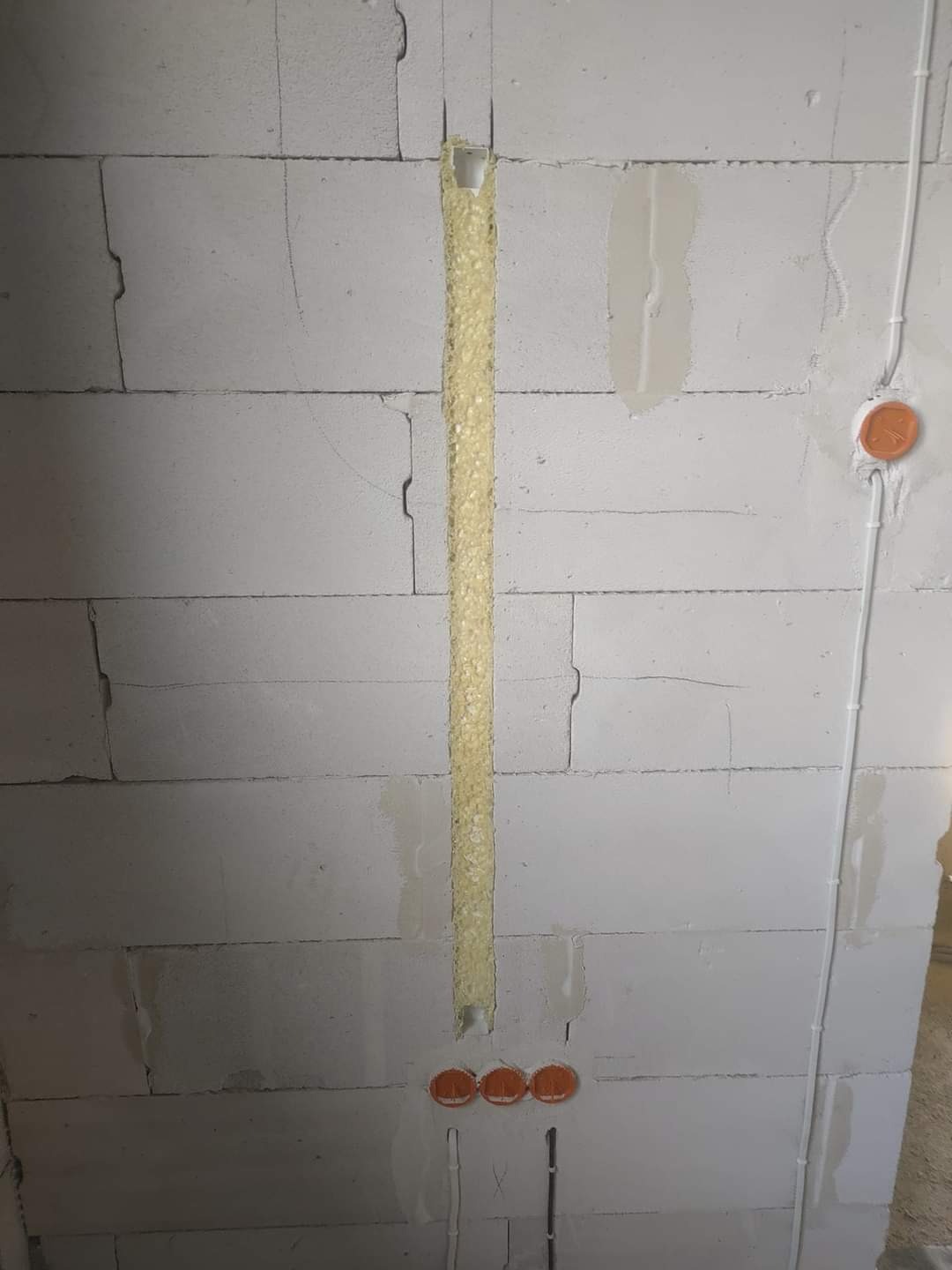 Brukowana ściana z pustaków z wyciętym pionowym rowkiem wypełnionym pianką montażową, poniżej widoczne trzy pomarańczowe puszki elektryczne, a po prawej stronie pionowy kabel elektryczny w białej...