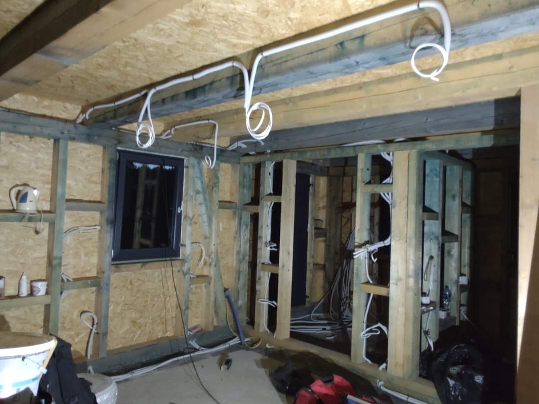 Wnętrze w trakcie budowy z widoczną drewnianą konstrukcją ścian i stropu, na których zamontowano białe przewody elektryczne prowadzone po powierzchni, tworzące pętle. Widoczne okno w czarnej ramie...