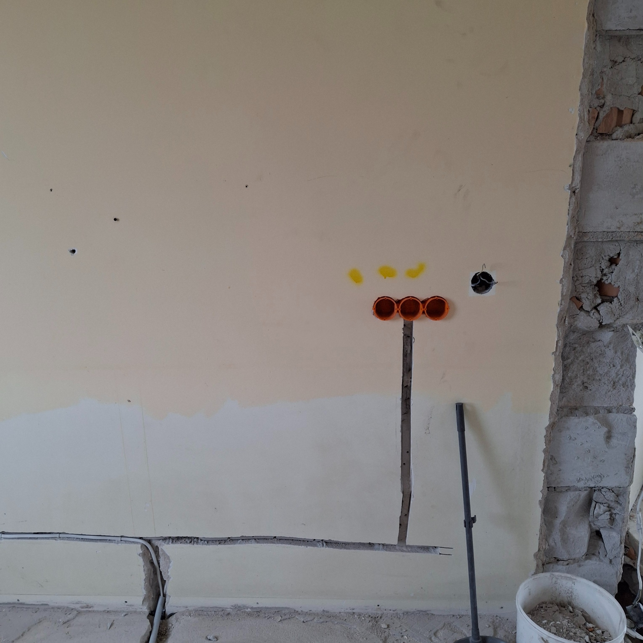 Ściana w trakcie remontu z widocznymi puszkami instalacyjnymi pod gniazdka i wyłączniki oraz bruzdami pod przewody elektryczne, obok fragment surowej ściany z cegły.