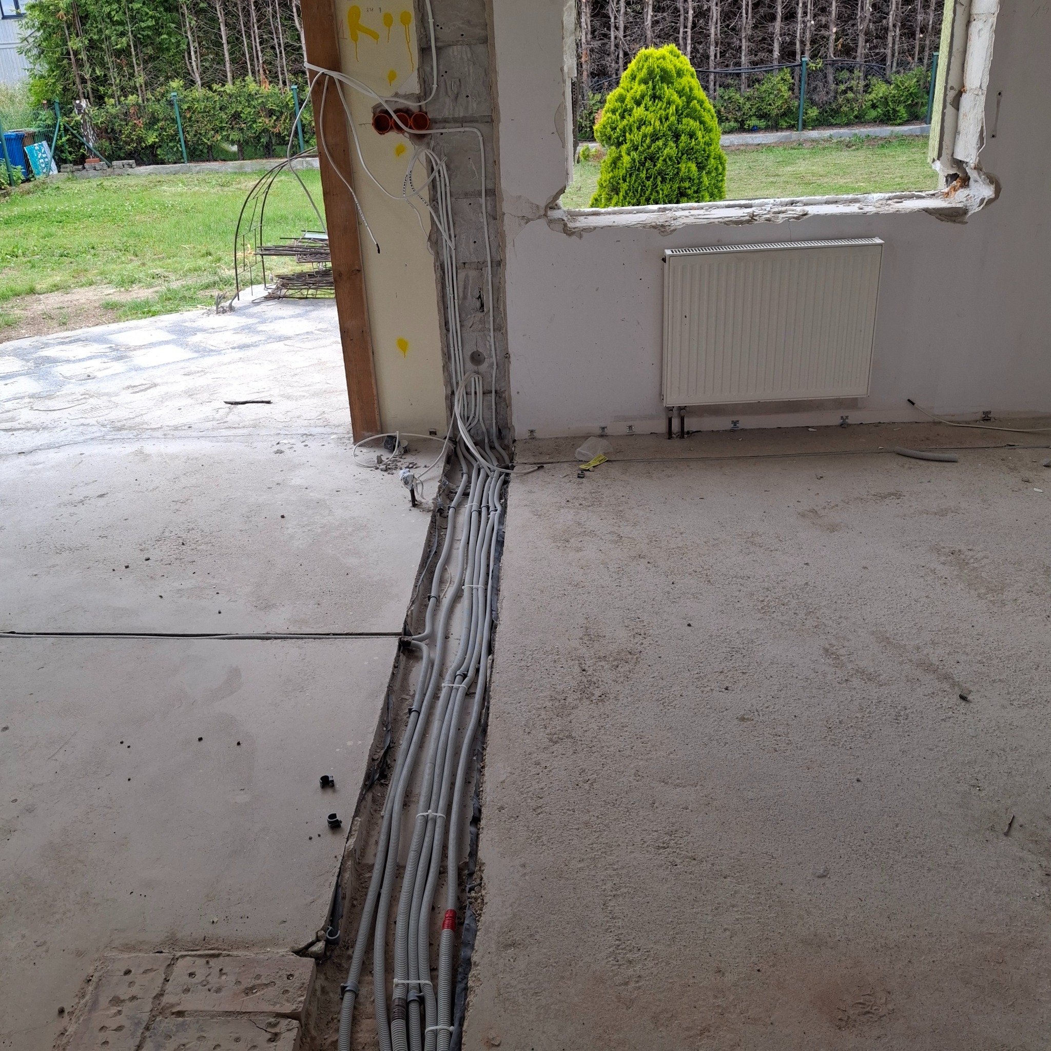 Szare kable elektryczne prowadzone w bruździe w ścianie, widoczne puszki instalacyjne i fragment widoku na zewnątrz przez okno w surowym stanie.