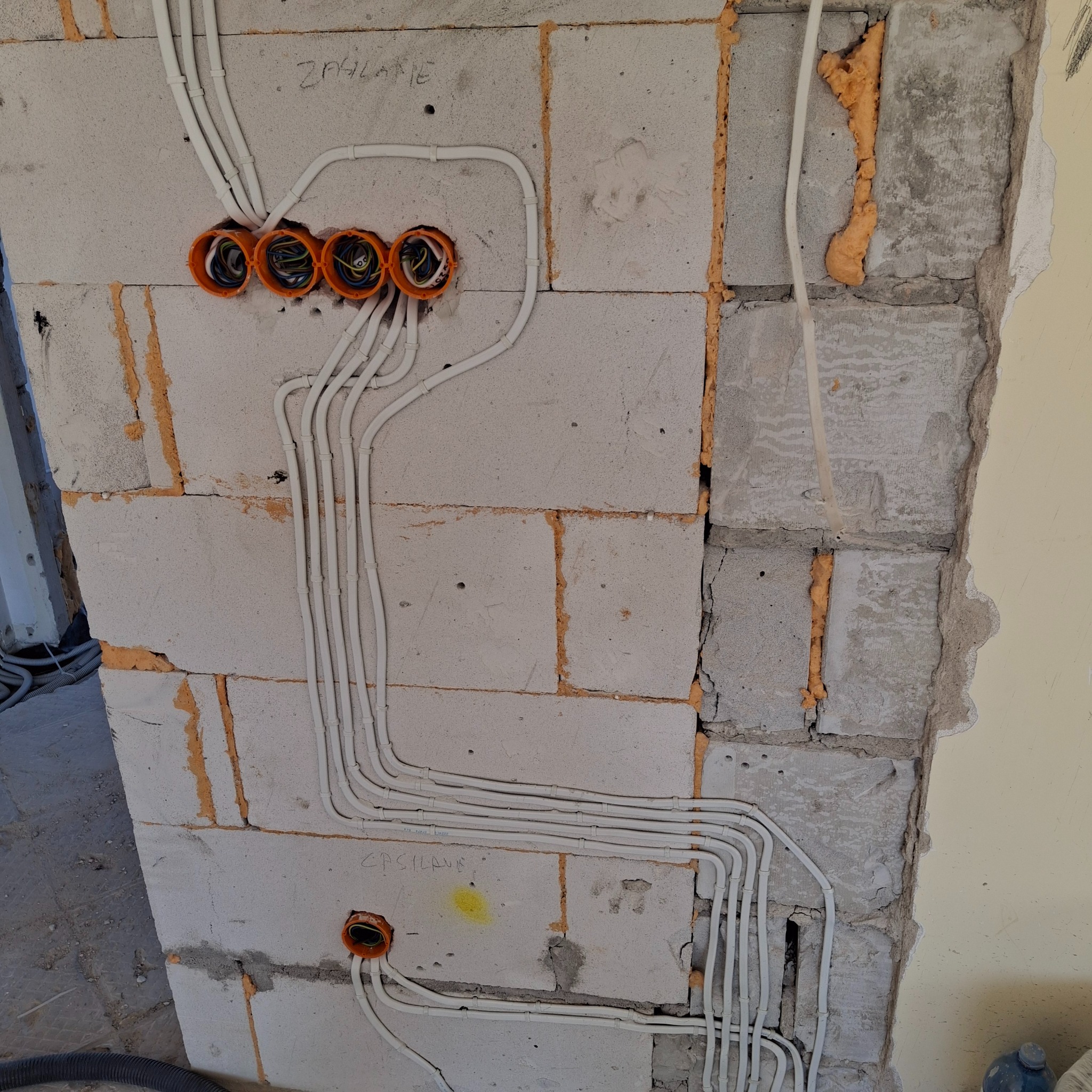 Białe przewody elektryczne ułożone w bruzdach ściany z betonu komórkowego, prowadzące do pomarańczowych puszek instalacyjnych, wypełnione pianką montażową szczeliny między blokami.