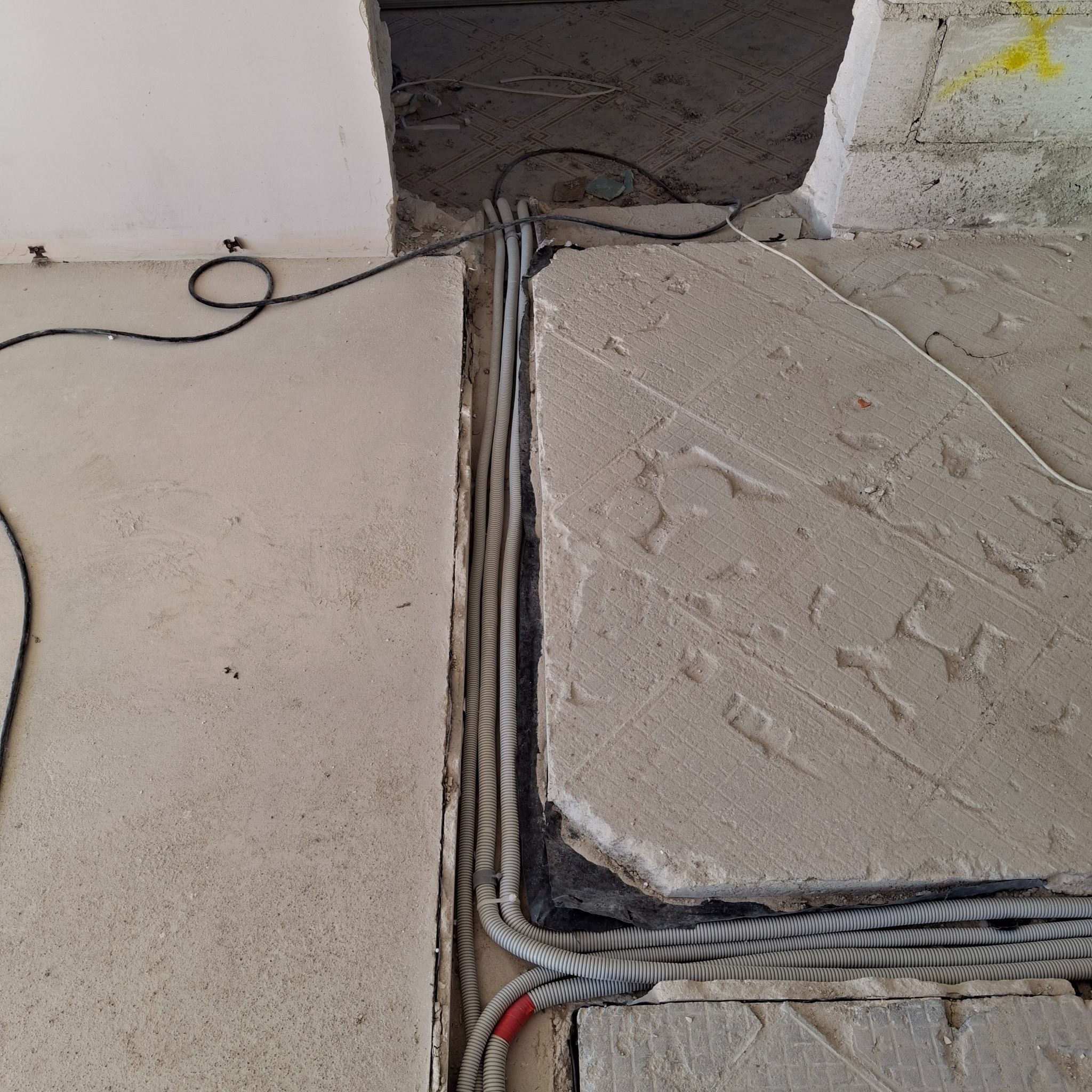 Kable elektryczne w szarych karbowanych rurach ułożone w bruździe w podłodze podczas remontu, widoczne fragmenty starej podłogi i ściany w surowym stanie.