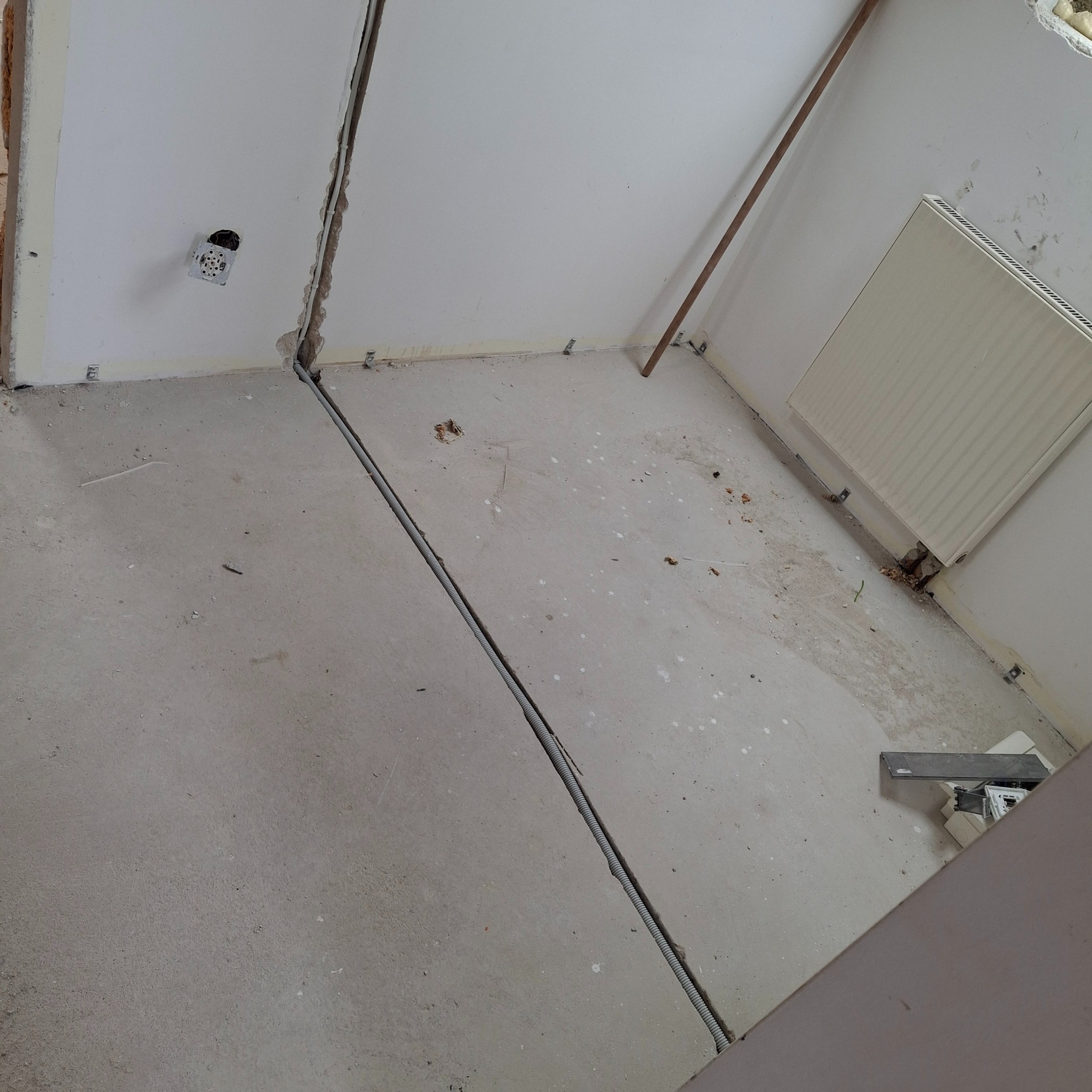 Szara rura karbowana w bruździe w podłodze, biegnąca od ściany do ściany, z widoczną puszką elektryczną w ścianie, przed remontem.