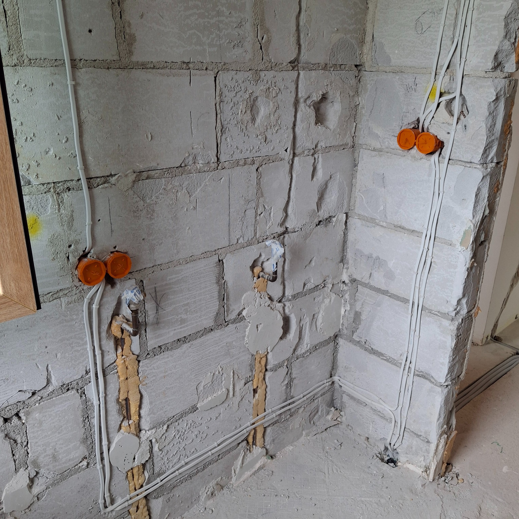 Surowe ściany z betonu komórkowego w trakcie prac. Widoczne pomarańczowe puszki elektryczne, białe przewody poprowadzone po ścianach i w bruzdach, częściowo zamaskowane pianką montażową i gipsem.