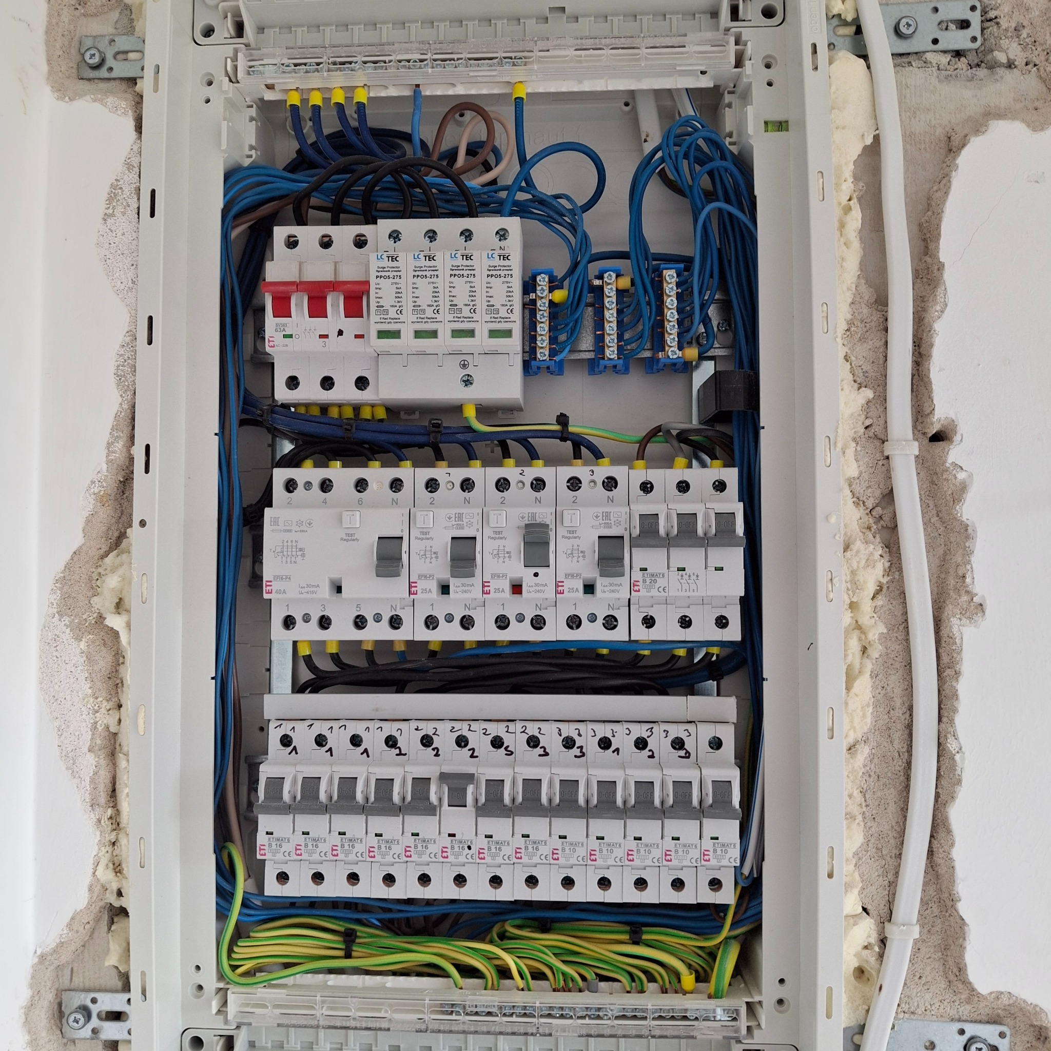 Rozdzielnica elektryczna w trakcie montażu, widoczne zabezpieczenia przepięciowe, wyłączniki nadprądowe, przewody w kolorach niebieskim, żółto-zielonym i czarnym, montaż w ścianie z pianką montażową.