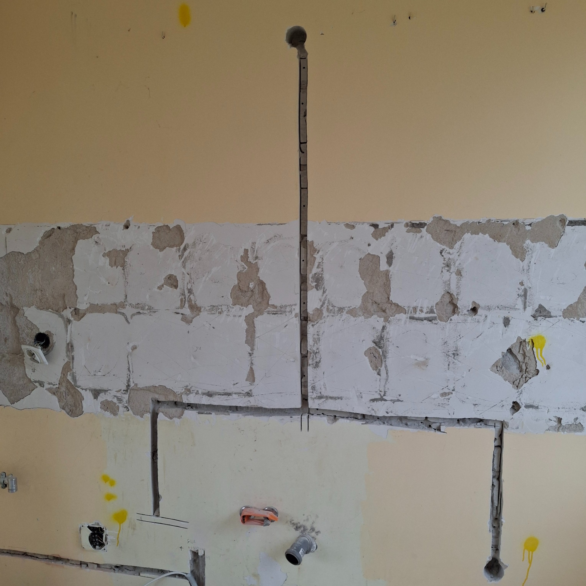 Przygotowana ściana z wyżłobieniami pod przewody elektryczne i gniazdka, widoczne ślady po kuciu, częściowo odkryta cegła, żółte plamy farby na ścianie.