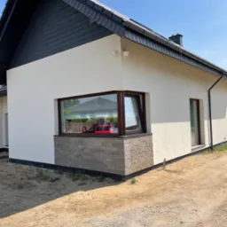 Wykończona elewacja domu jednorodzinnego z białym tynkiem i kamienną okładziną pod oknem, widoczne okna z brązowymi ramami i czarny dach z szarymi rynnami, na pierwszym planie piaszczysty teren.
