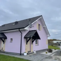 Fioletowy dom jednorodzinny z czarnym dachem i zadaszeniami nad wejściami, widok z zewnątrz na szarym brukowanym podjeździe i trawniku.