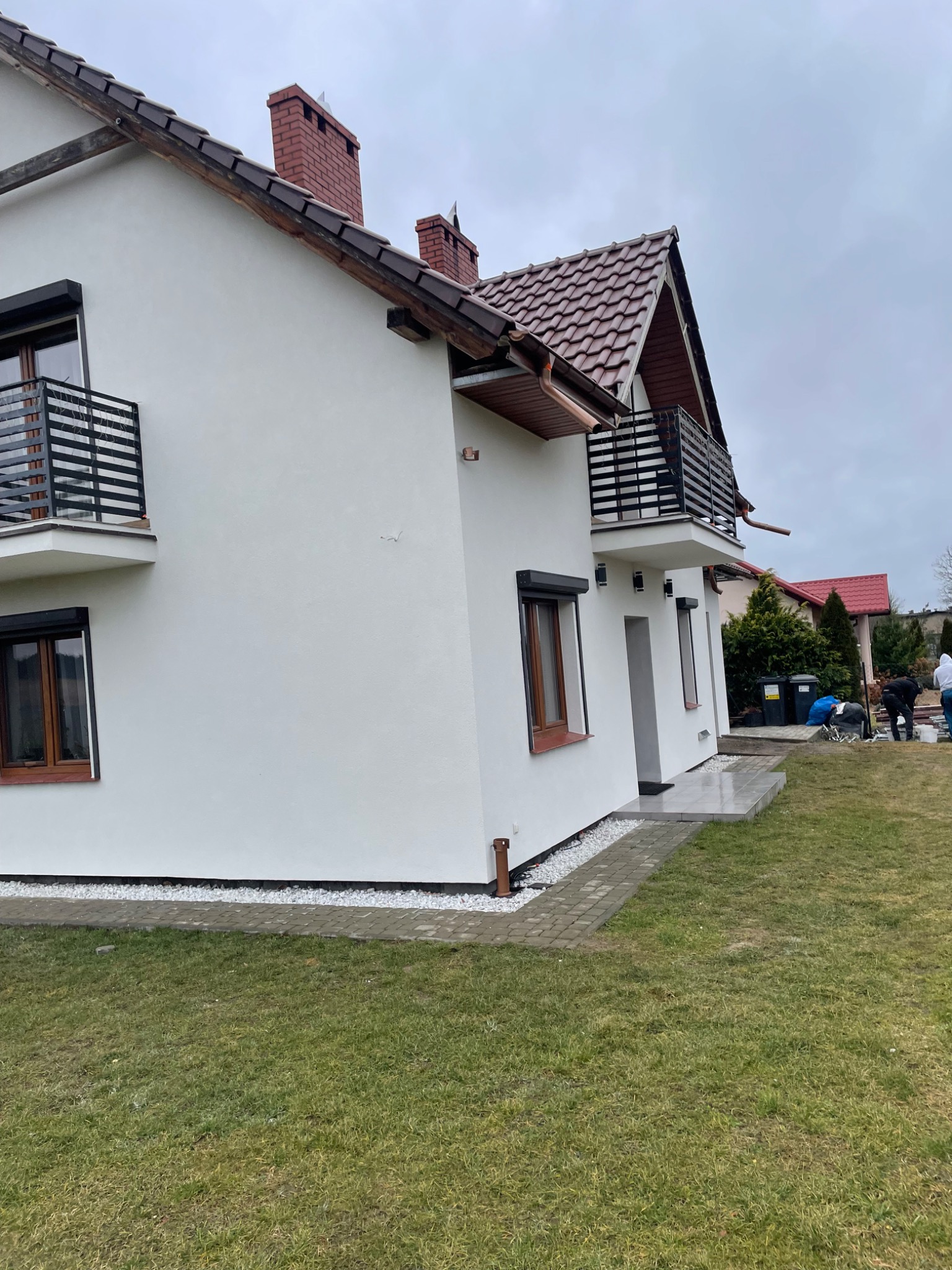 Świeżo otynkowany i pomalowany na biało dom jednorodzinny z balkonami i brązowymi oknami, widoczny z perspektywy ogrodu, z dwoma osobami pracującymi w tle.