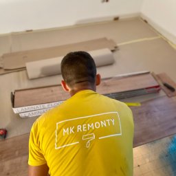 Kompleksowe Wykończenia Wnętrz - Pracownik w żółtej koszulce z logo firmy 'MK Remonty' układa panele podłogowe w pokoju, widoczne paczki paneli i narzędzia