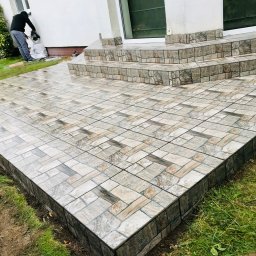 Kompleksowe Wykończenia Wnętrz - Wyłożony płytkami taras z niskimi schodami wejściowymi, widoczna osoba sprzątająca po pracy.