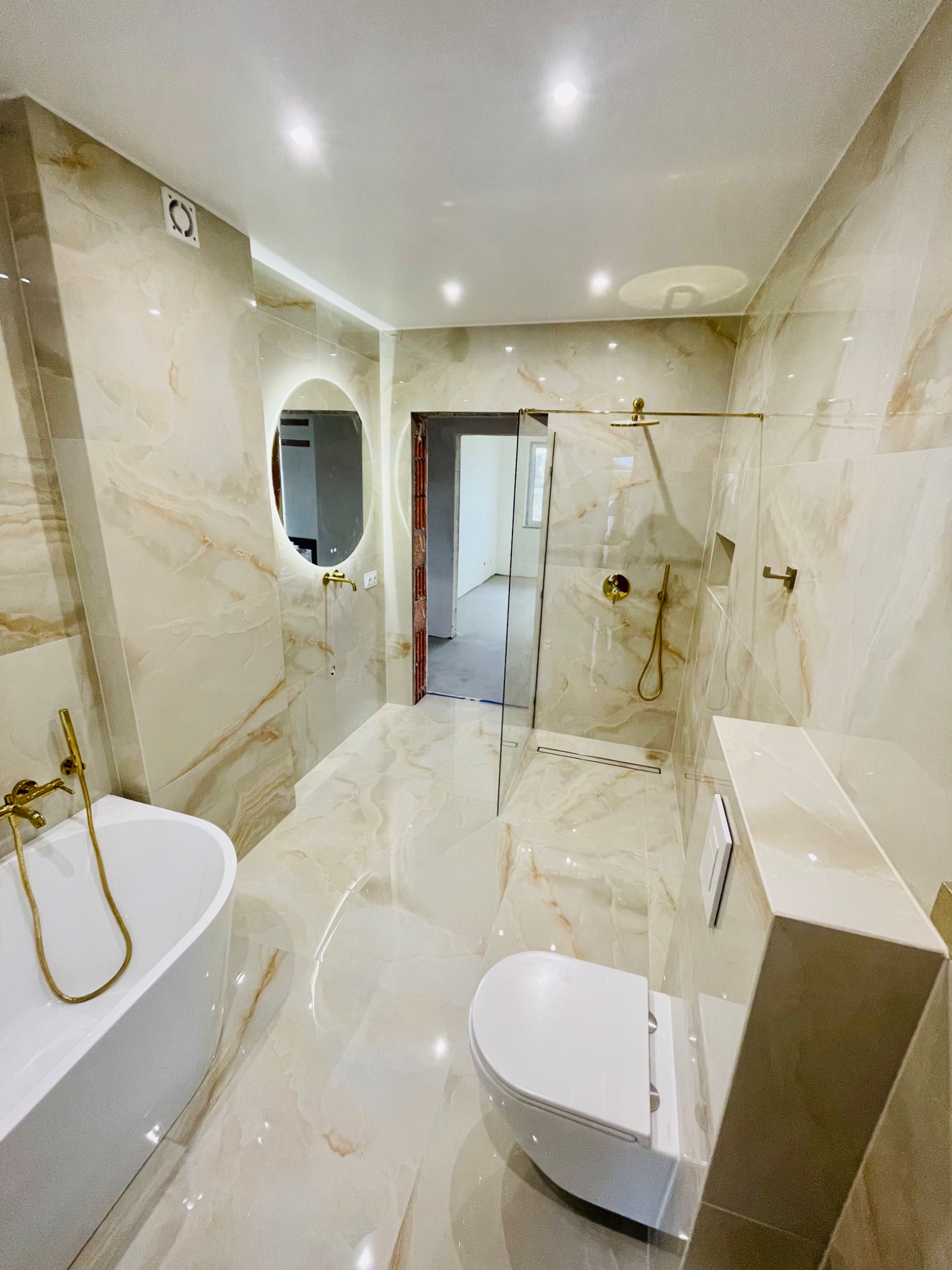 Nowoczesna łazienka wykończona płytkami imitującymi marmur, z kabiną prysznicową typu walk-in ze złotymi okuciami, owalnym podświetlanym lustrem i białą wolnostojącą wanną.