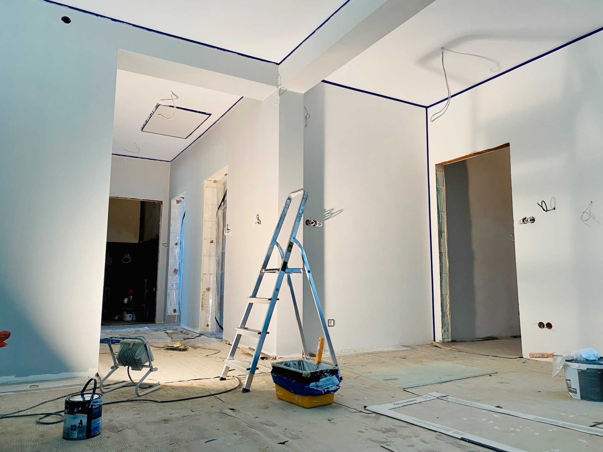 Wnętrze w trakcie odświeżania, z pomalowanymi na biało ścianami i niebieskimi liniami wyznaczającymi granice, drabina malarska, wiadro z farbą, lampa halogenowa na podłodze zabezpieczonej folią...