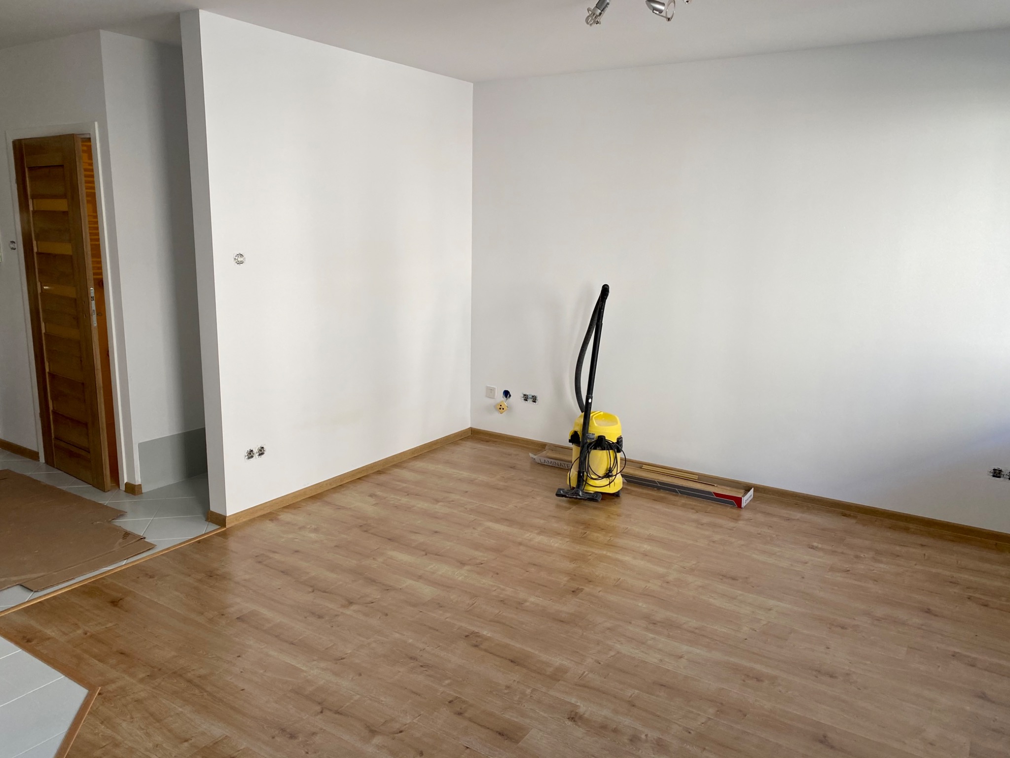 Pokój po remoncie z nową podłogą z paneli laminowanych, widoczny odkurzacz, listwy przypodłogowe i fragment płytek w przejściu do innego pomieszczenia.