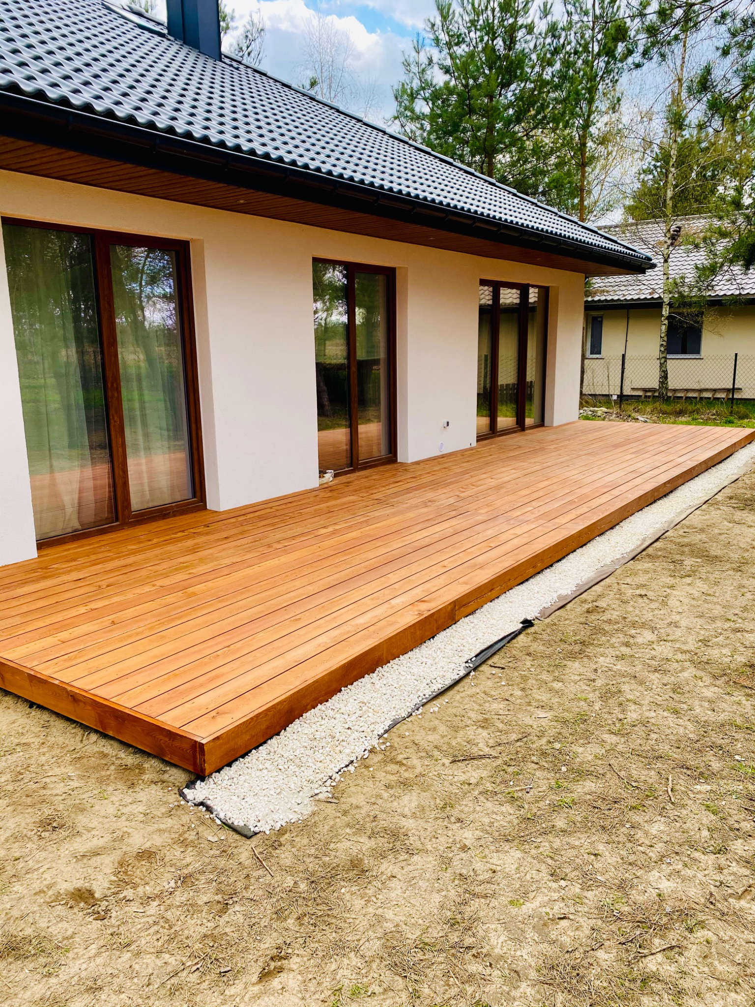 Nowoczesny taras z desek kompozytowych w odcieniach brązu, przylegający do jasnej elewacji domu z ciemną dachówką, obrzeże z białego grysu oddziela taras od trawnika.